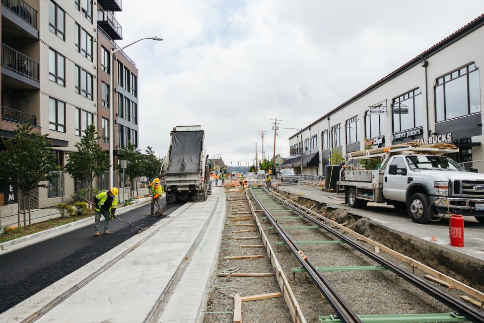 Buildings line a wide street, and tracks for the Hilltop Tacoma Link Extension are being laid in the middle of the road.