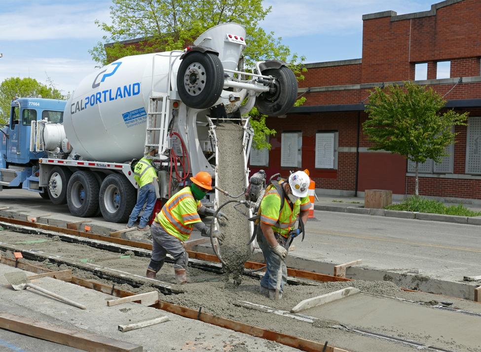 Two workers in vests and hard hats use a machine to pour concrete in Tacoma for a new rail extension.