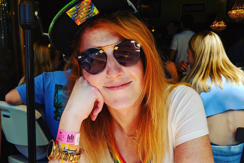 Mattie Tharpe wears sunglasses, a baseball hat and a T-shirt with rainbow trim. She rests her face on one hand while smiling at the camera, sitting at a picnic table.