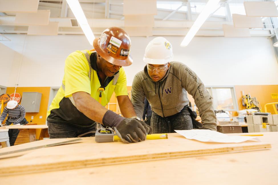 Two men in hard hats reach over a drafting table to work on a construction project inside a classroom-type setting.