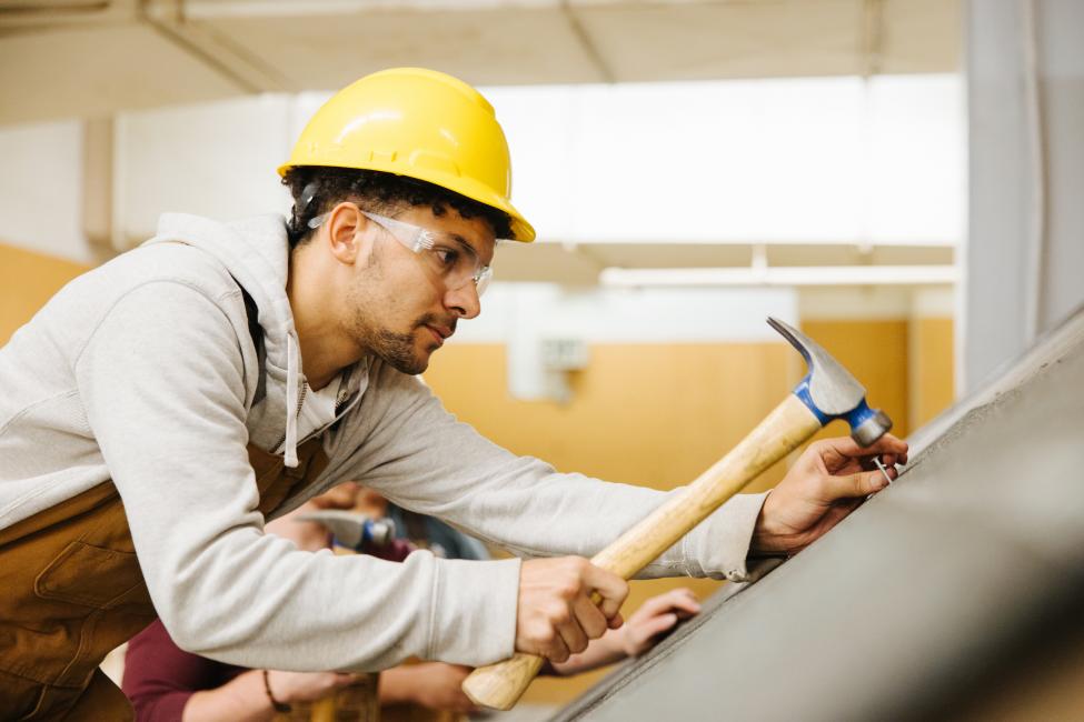 A young man wearing overalls an a hard hat holds a hammer while learning roofing skills.