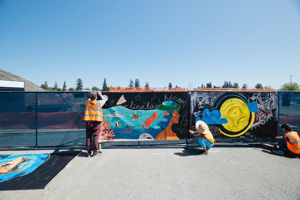 Three Sound Transit employees in orange vests install colorful murals on blue construction fencing.