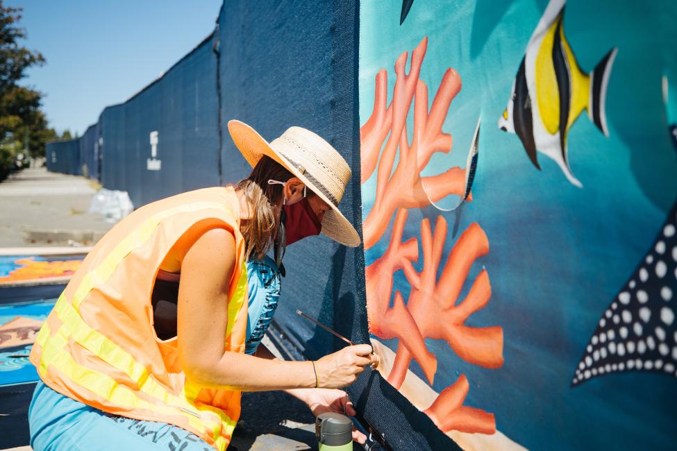 A woman in an orange vest and sun hat attaches a colorful mural depicting coral and fish to a construction fence.