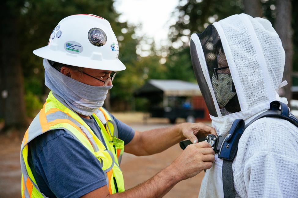 A man in a hard hat, mask and yellow vest outfits another man in a white beekeeper's suit.