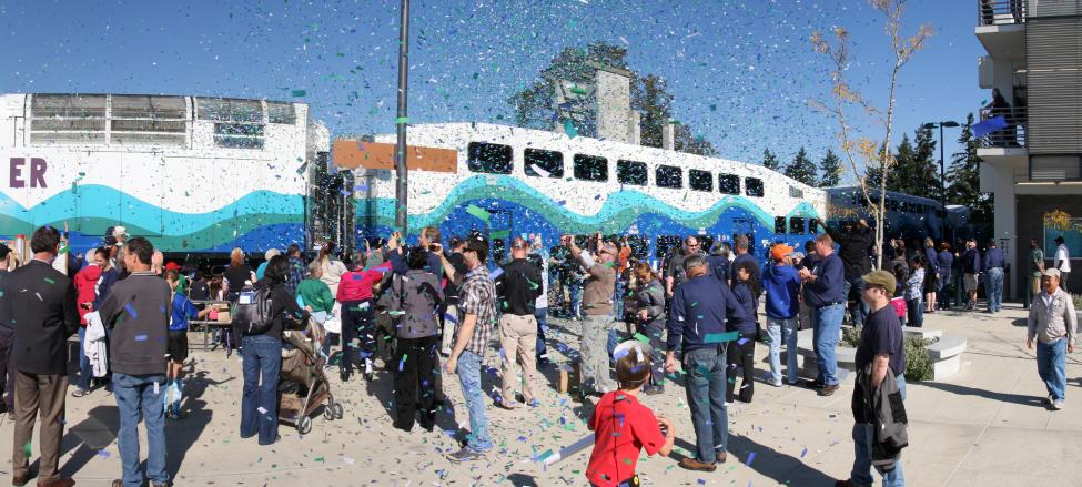 Confetti flies as a crowd stands in front of a Sounder train (white with a blue and green wave on the side).