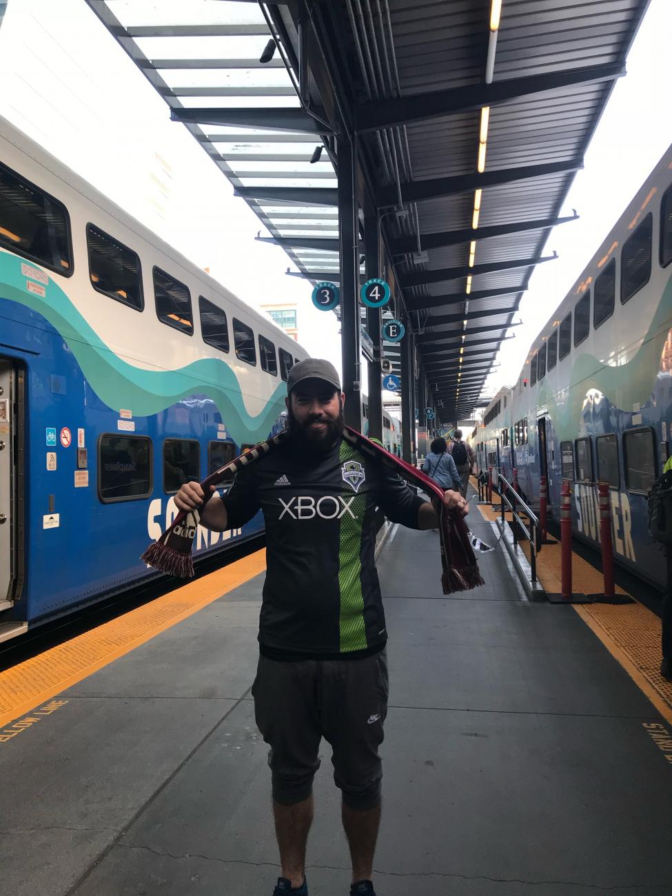 A man stands on the Sounder train platform, wearing a Sounders soccer team jersey and scarf.
