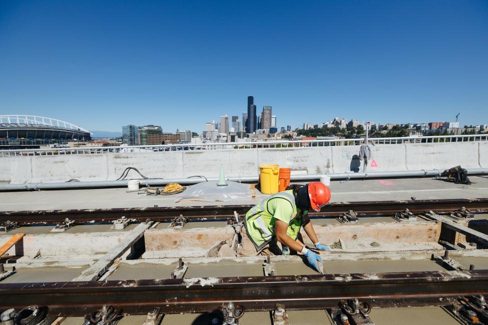 A worker in a yellow vest and orange hard hat kneels by new light rail tracks, with the downtown Seattle skyline in the background.