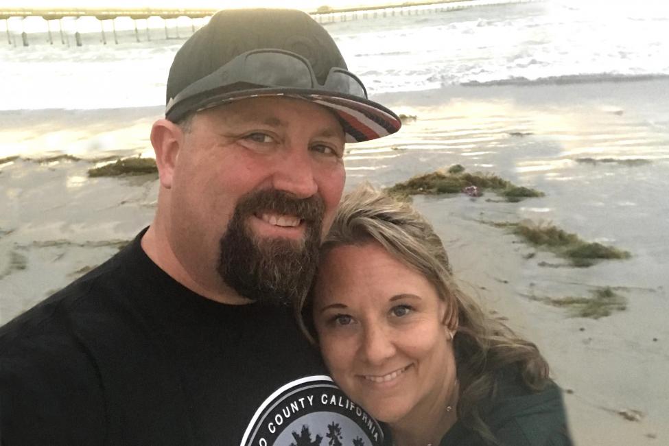 Rob McMartin - a man wearing a black shirt and green baseball hat - and his wife smile while taking a selfie on the beach.