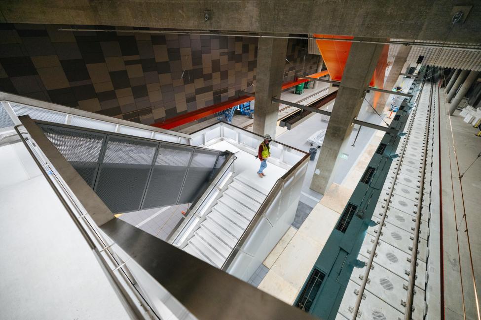 A construction worker walks down a staircase to the platform of University District Station. Train tracks are visible, and the station is well lit.