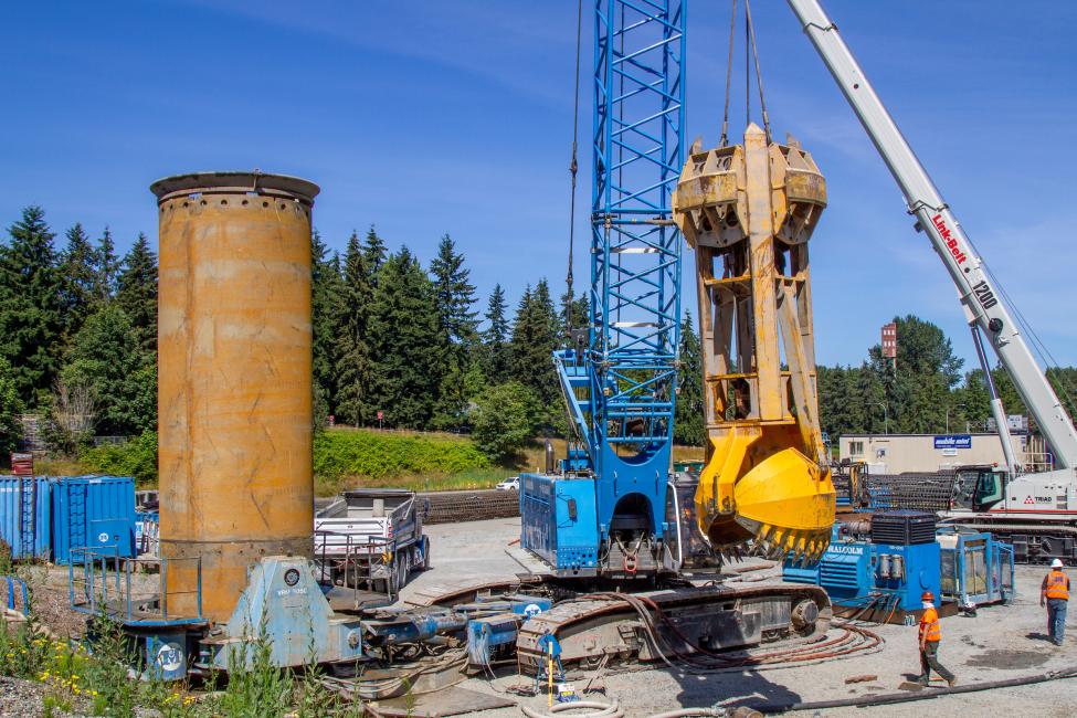 A construction worker in the bottom right corner of the photo is dwarfed by construction equipment and a large drill shaft.