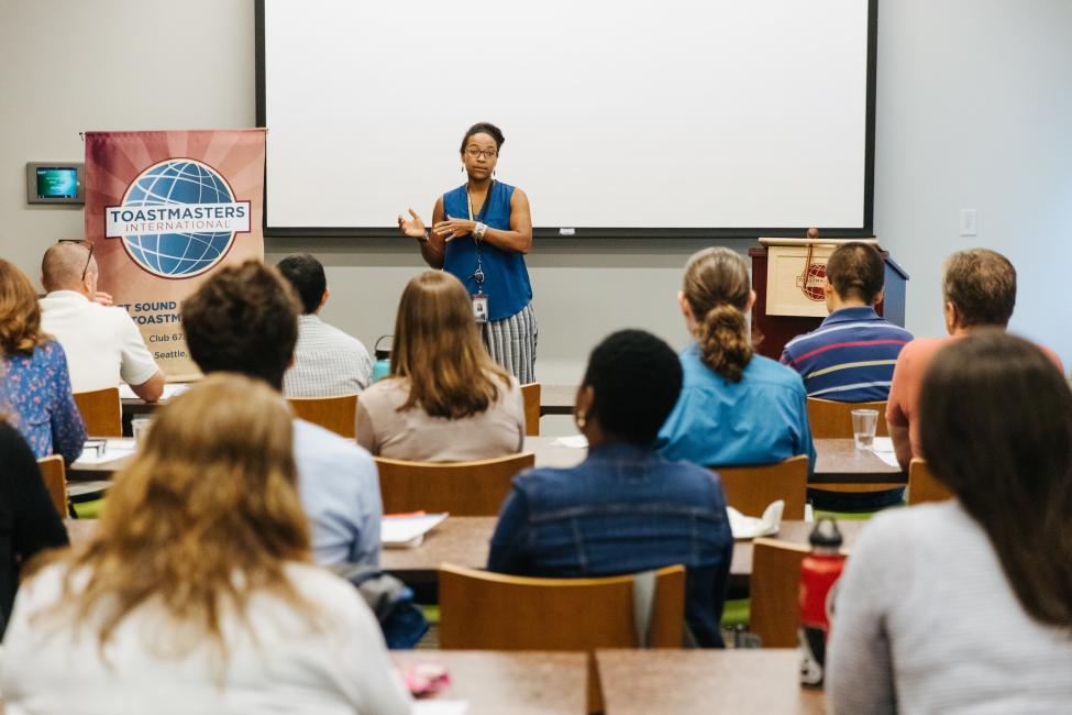 Andrea Stuart-Lehalle speaks to a crowd at a Toastmasters training, held in an indoor room. A projector screen is behind her.