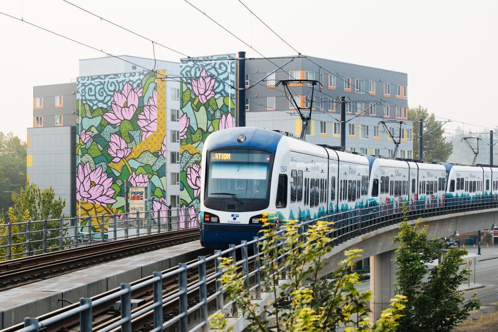 A Link train travels along an elevated track in the Mt. Baker neighborhood, with a colorful mural of flowers visible on the side of a building in the background.