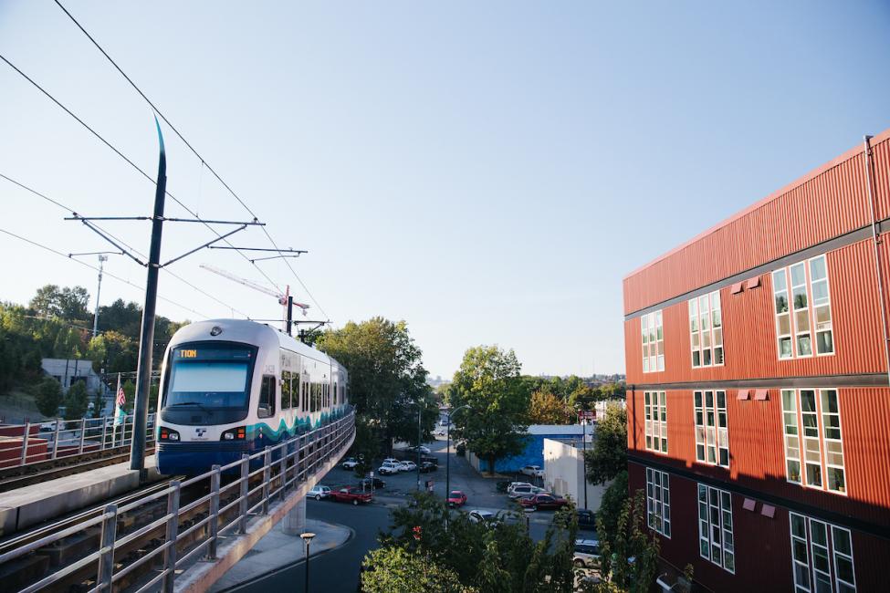 A Link train travels along elevated tracks to the left of a large brick building.