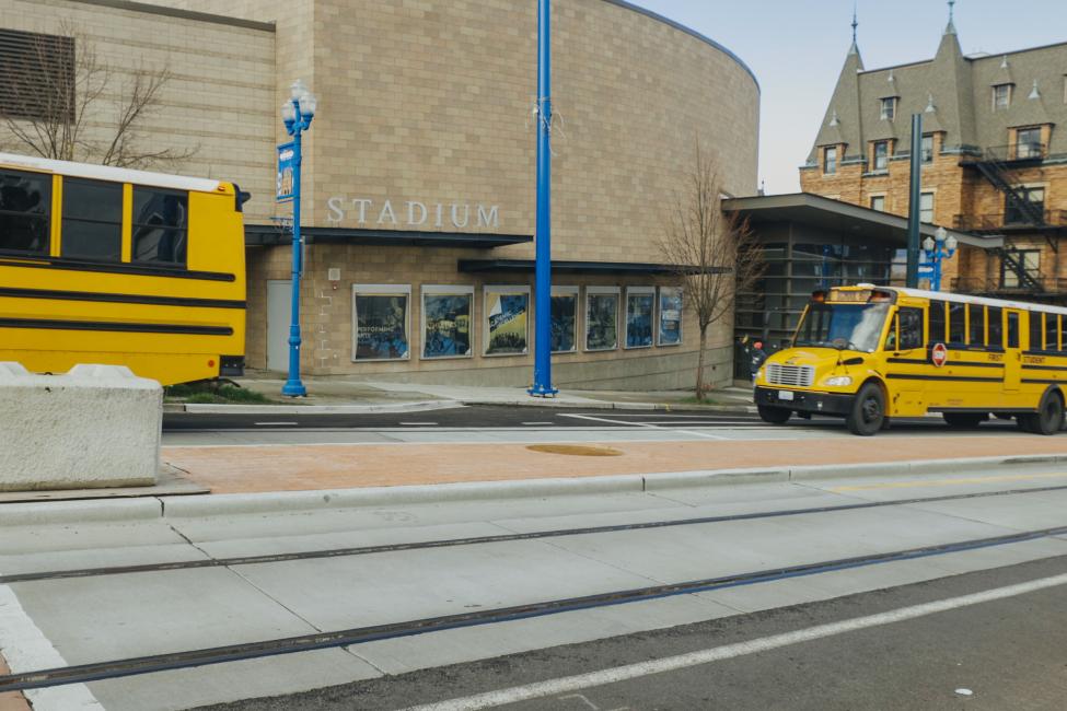 Two yellow school buses are parked by a stadium, with new light rail tracks on the street in the foreground.
