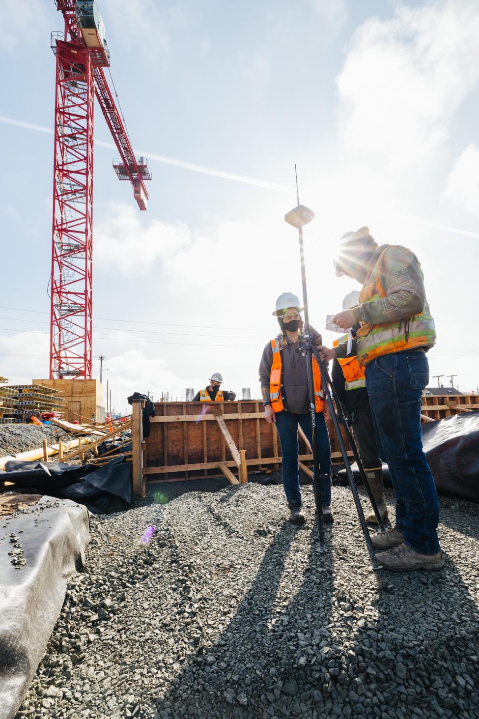 The sun shines on three construction workers as they look down at a tablet while doing survey work.