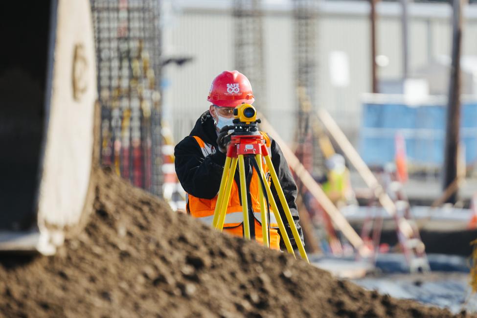A construction worker in a red hard hat does survey work by looking through a piece of equipment on a tripod.