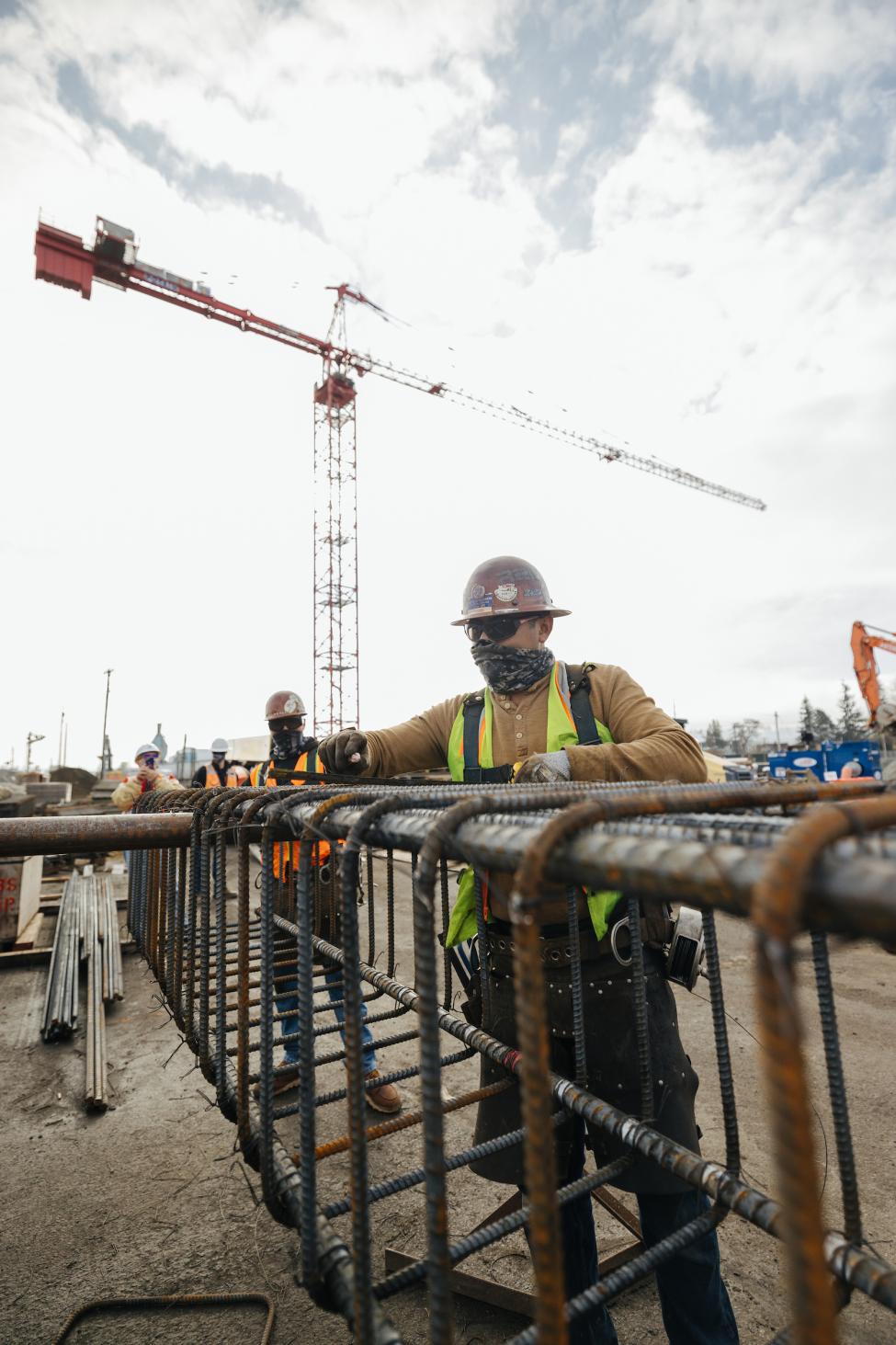 Workers wearing face coverings, hard hats and sunglasses reinforce steel on a rebar cage.