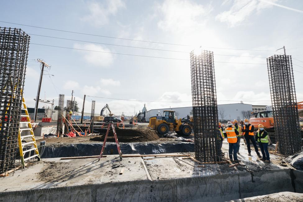 Members of a construction crew wearing vests and hard hats stand to the right of three cages made of rebar.