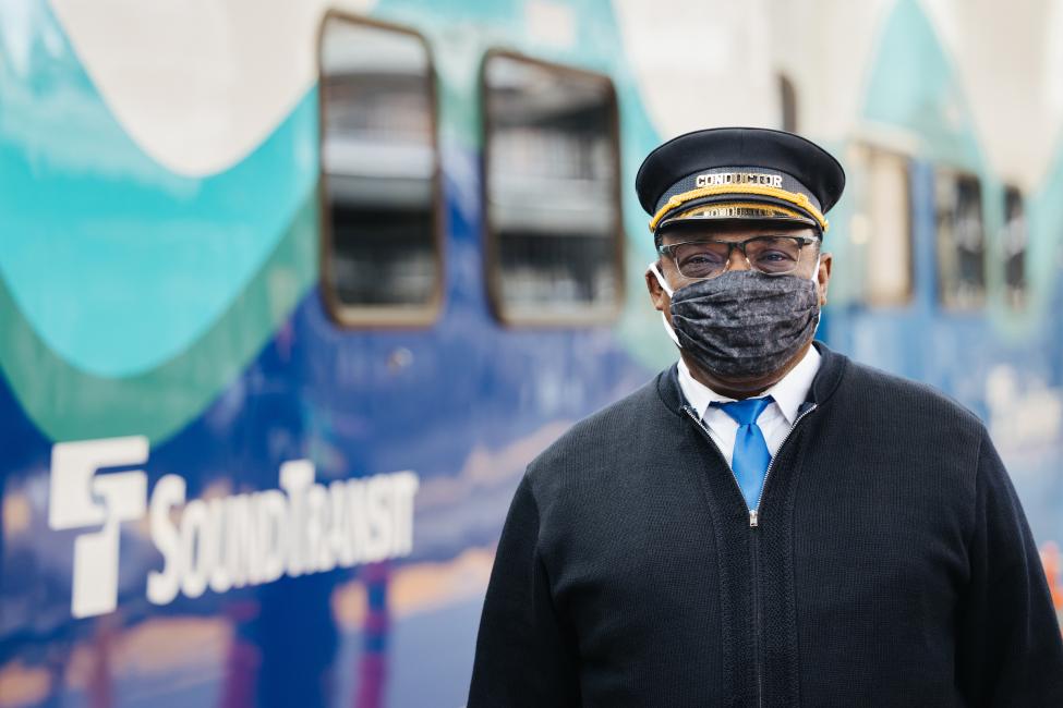 A man wearing a mask and conductor hat stands next to a Sounder train.