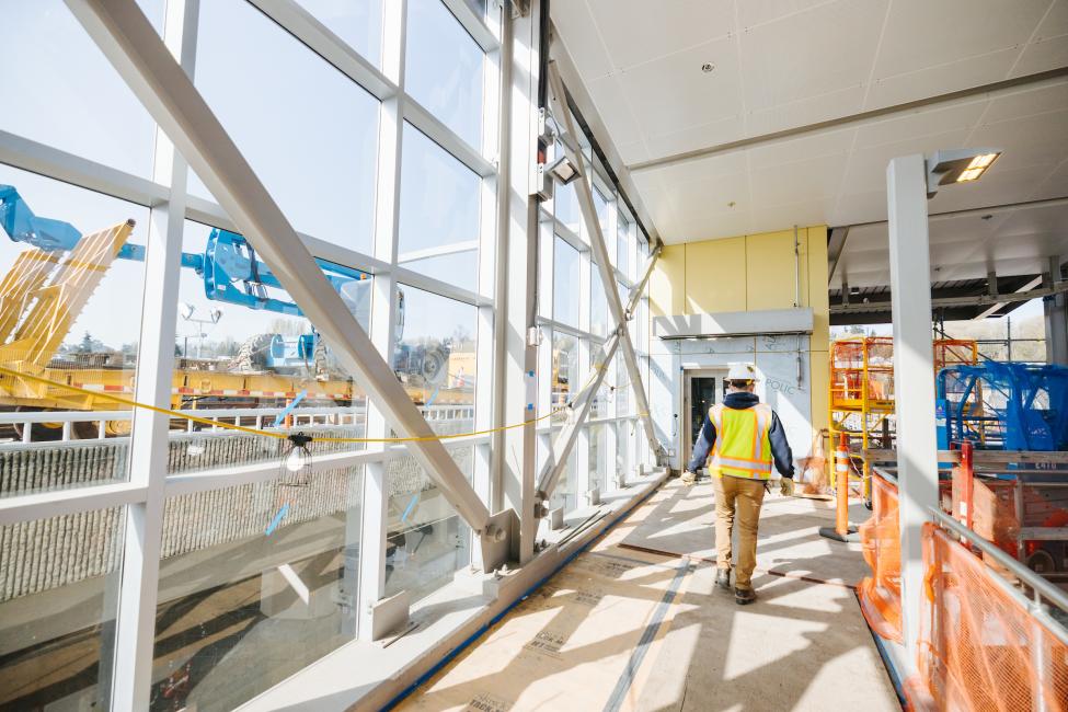 A construction worker walks away from the camera next to a glass wall at Judkins Park Station on a sunny day.