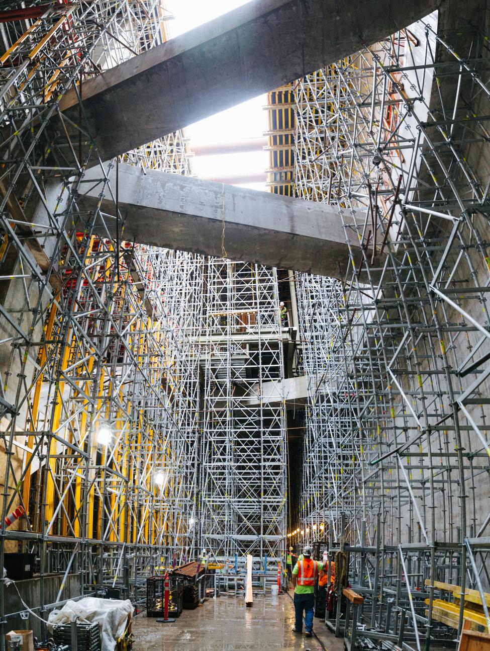 Two people in orange vests look up at giant pieces of steel falsework at the U District light rail station while it was being constructed.