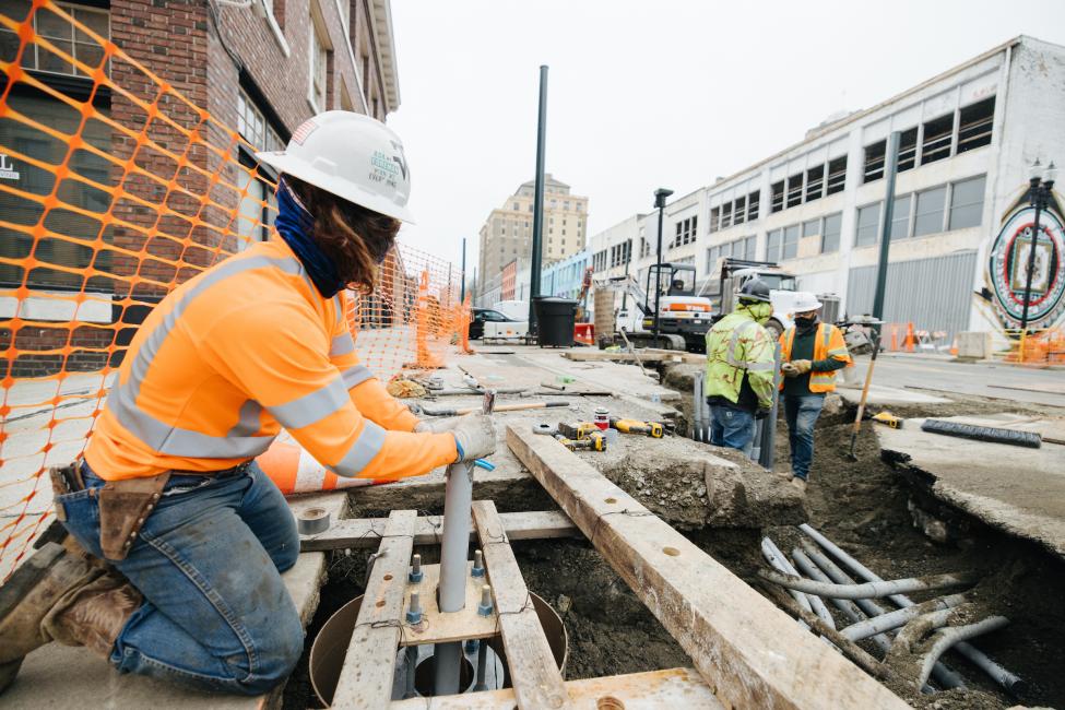 A worker in an orange jacket and white hard hat does utility work in the street as part of the Hilltop Tacoma Link Extension.