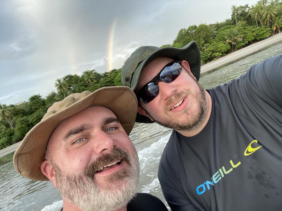 Two men take a selfie on a boat. Both are wearing hats and one is wearing sunglasses.
