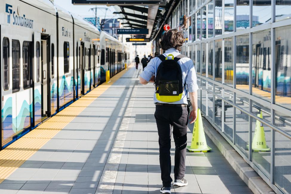 A Link passenger walks along a train at the Mount Baker Station platform. He is wearing a black backpack and walking away from the camera.
