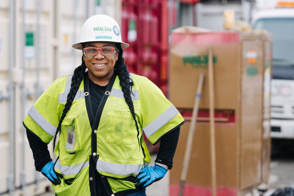 Andrea Ornelas smiles while wearing a white hard hat, red glasses and a yellow jacket.