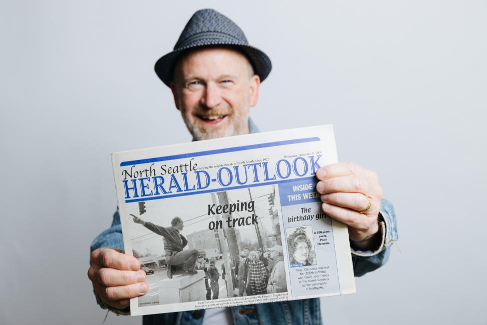 Photo of Jim O'Halloran holding the North Seattle Herald-Outlook newspaper from 2004 that features a front page photo of him leading a tour of the area where his organization lobbied successfully to locate the Roosevelt Station. 