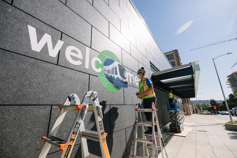 A worker stands on a ladder while installing a sign that says "Welcome" on the exterior of U District Station.