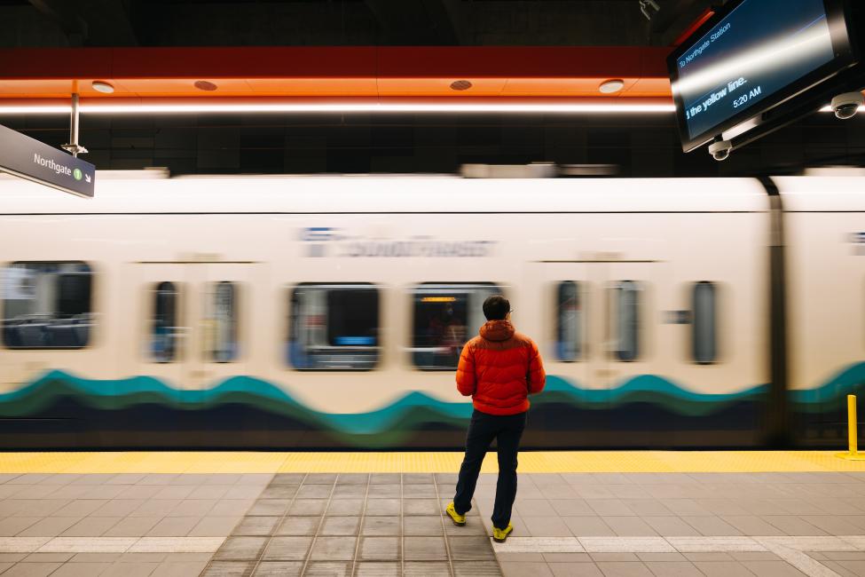 A man in an orange jacket watches a Link train zip by.