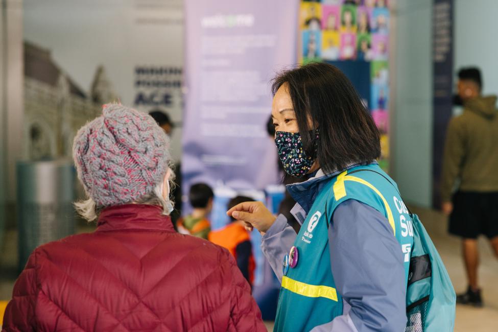 A woman in a mask and teal vest talks to a rider. 