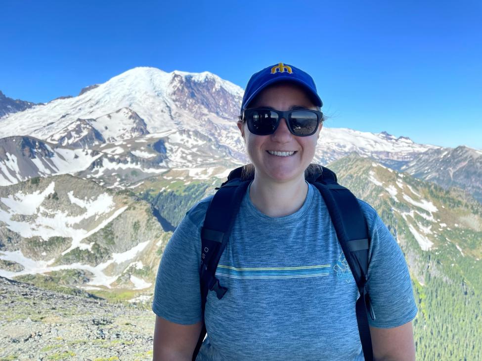 A woman in a baseball hat and sunglasses smiles with a mountain in the background.