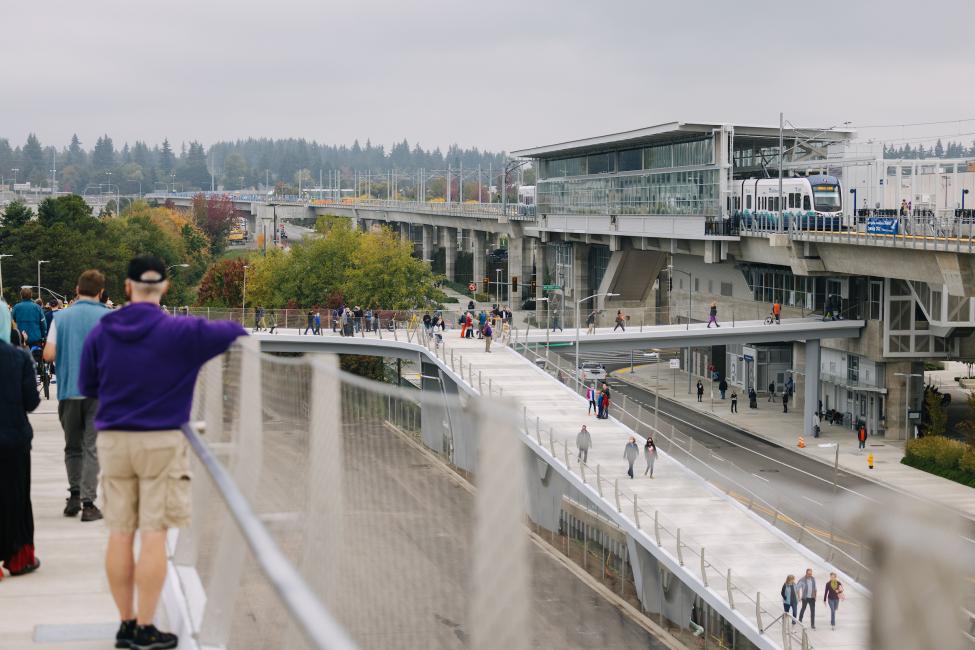 A view of Northgate Station on opening day from the new John Lewis Memorial Bridge for pedestrians that crosses over I-5 as people walk and push bikes over the bridge coming and going to/from the new station. 