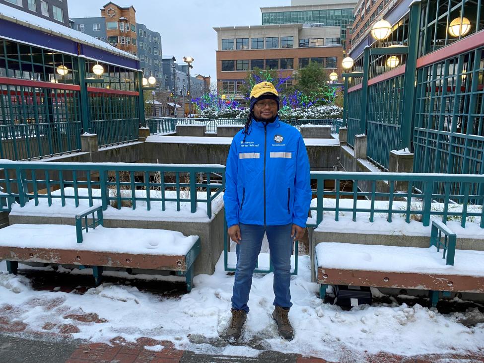 A fare ambassador wearing a blue vest and yellow cap stands outside a light rail station in the snow.