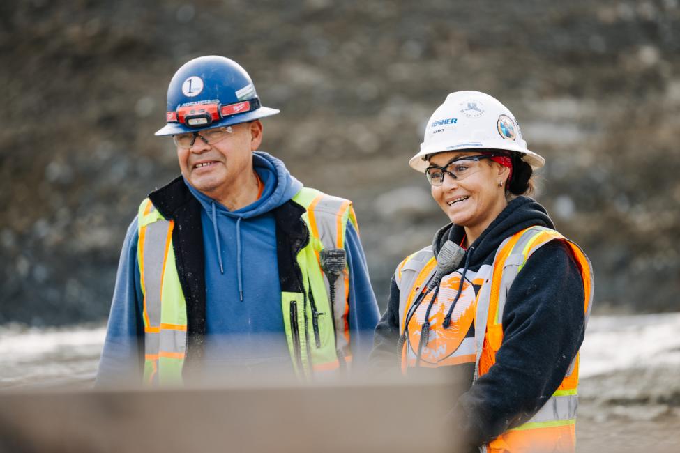 A man and a woman in construction gear smile while looking out over a work site. 
