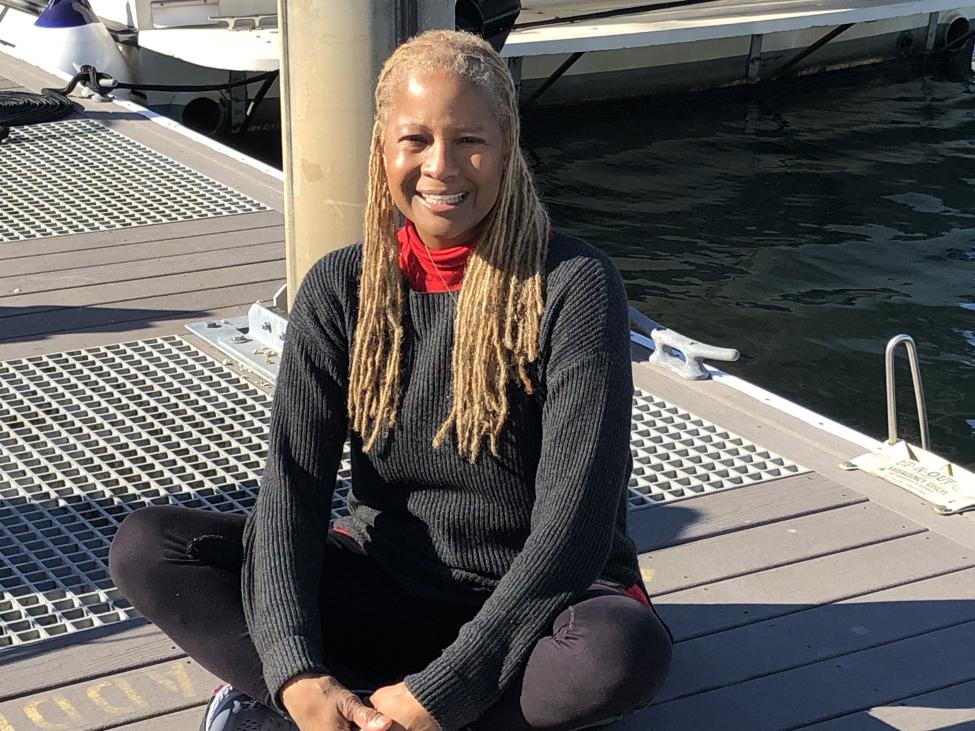 Galen sits cross-legged on a boat dock, wearing a gray sweater over a red turtleneck.