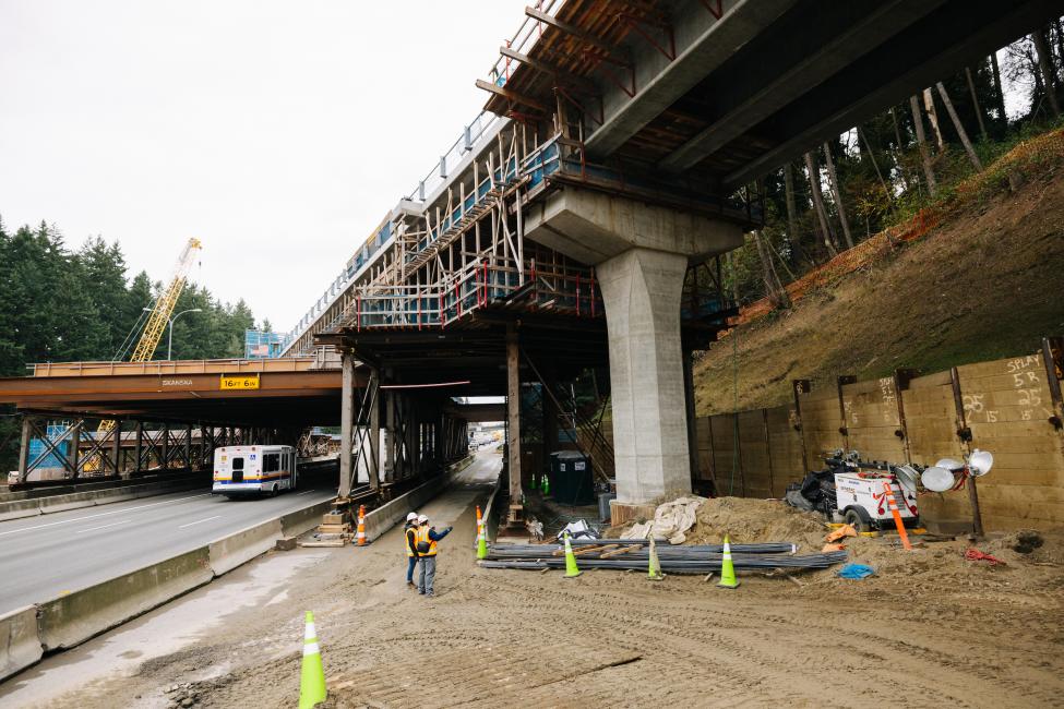 Two people in construction vests and hard hats look up at a bridge under construction.