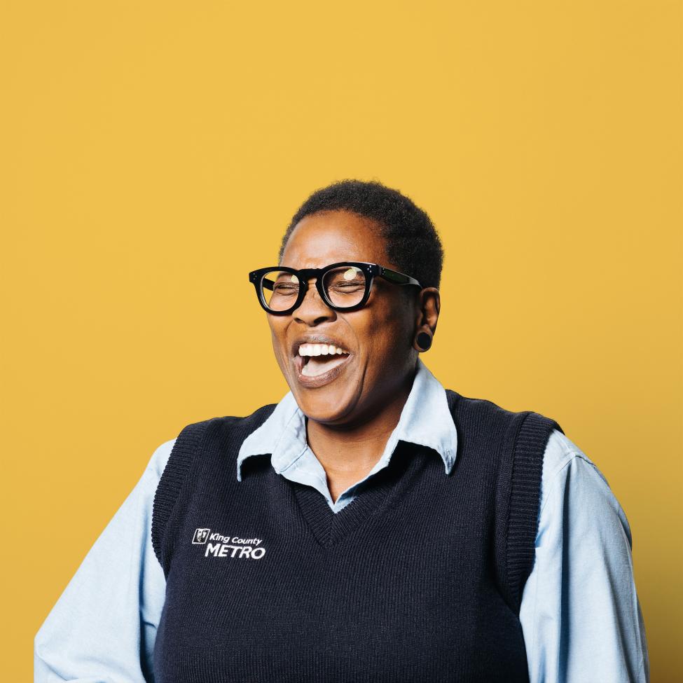 Donna Newton, a King County Metro operator, smiles in front of a yellow background. She wears chunky glasses and a sweater vest.