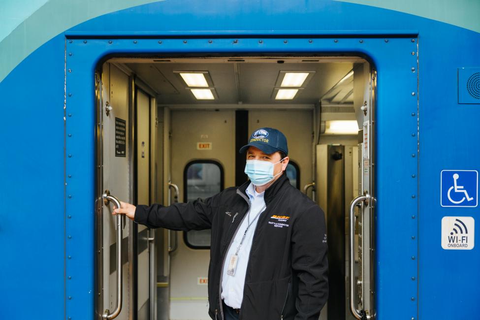 A Sounder conductor wears a hat and mask and smiles while posing near the train door.