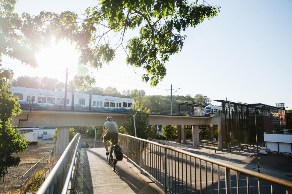 A Link light rail train approaches Mt. Baker Station in the background as a bike rider crosses a bridge over the road in the foreground.