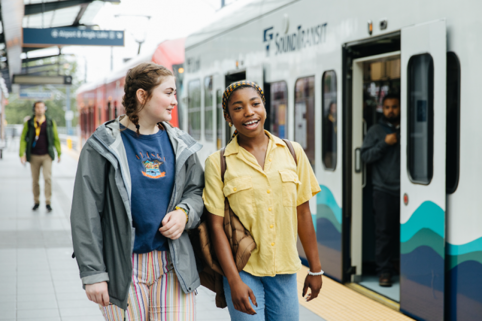 Two young people walk on a platform next to a Link train.