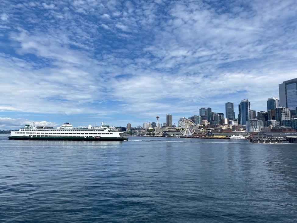 A ferry departs from the terminal in downtown Seattle. The skyline, including the Space Needle and Great Wheel, is in the background.
