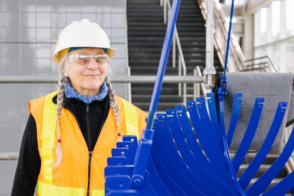 Artist Beliz Brother supervises the installation at Mercer Island Station. She wears an orange vest and white hard hat and looks at a blue metal sculpture of a rowboat. 