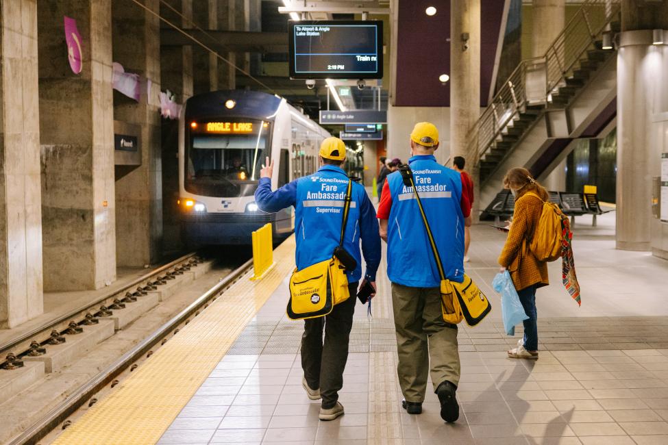 Two fare ambassadors walk away from the camera and toward a Link train at Roosevelt Station.