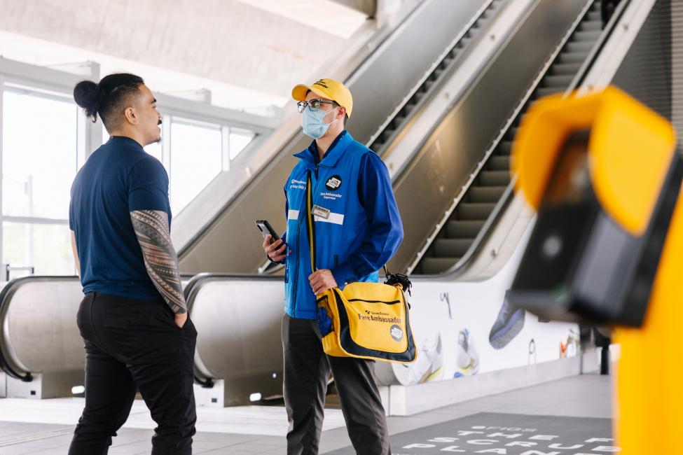 A fare ambassador in a face mask, yellow hat and blue vest talks to a passenger at Northgate Station.