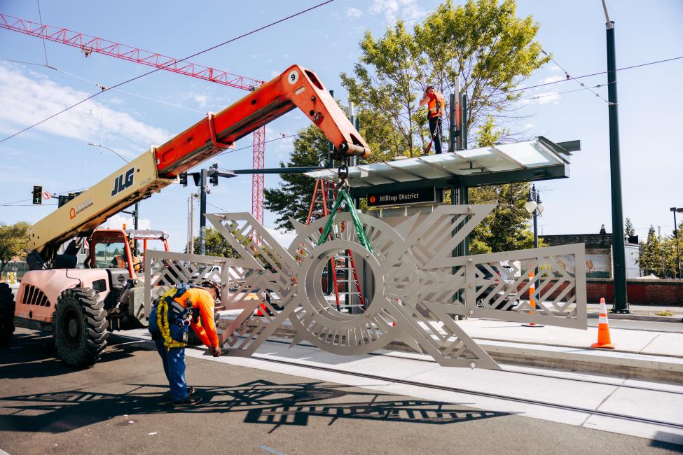 An art piece rests on the ground as construction crews get ready to use machinery to lift and install it.