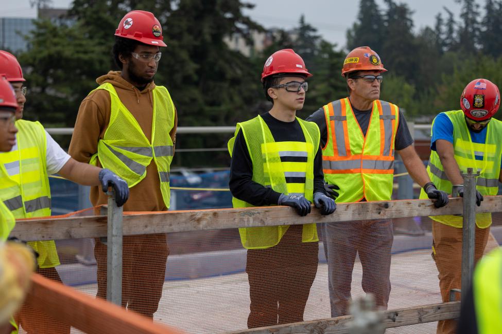 Five people wearing yellow vests and hard hats stand in a line on a construction site.