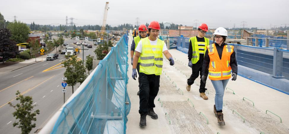 A group of people wearing protective gear walk along a light rail guideway under construction.